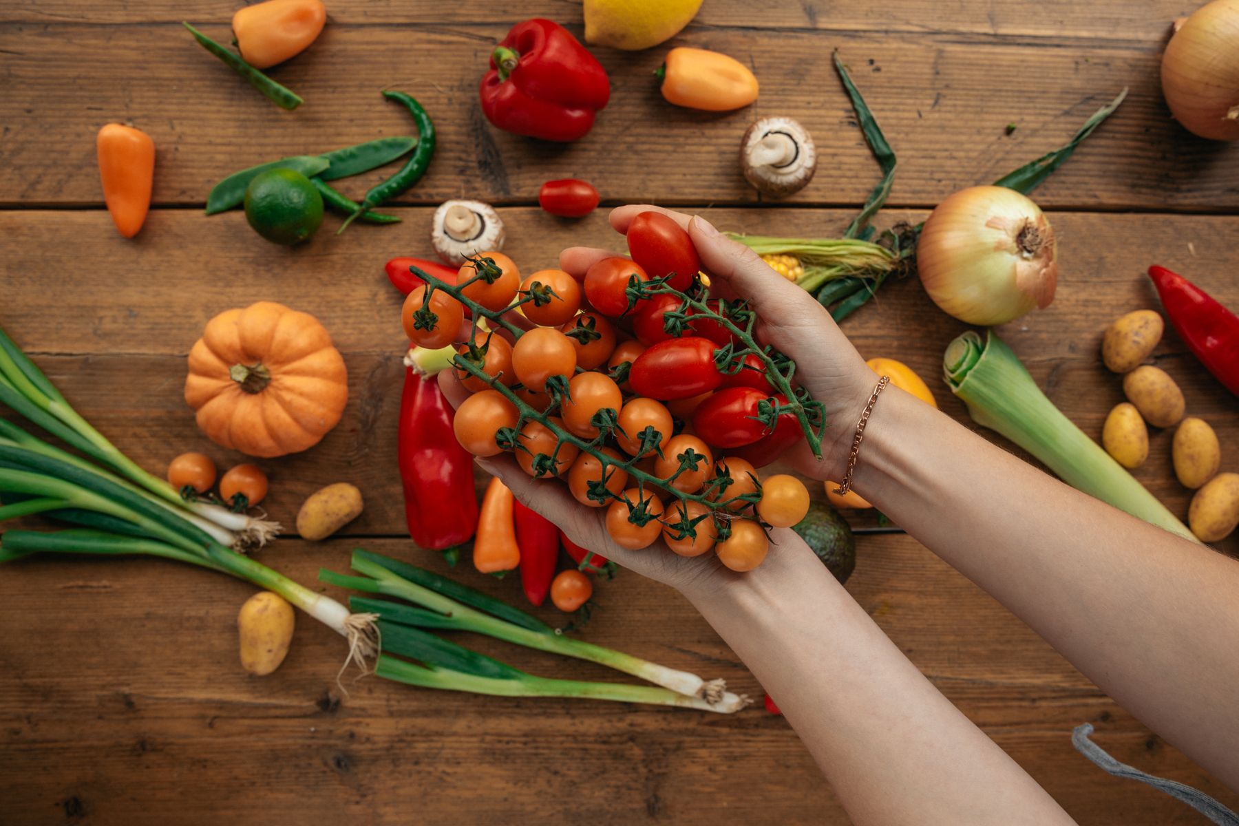 Hands cradling fresh cherry tomatoes over a wooden table spread with colorful vegetables